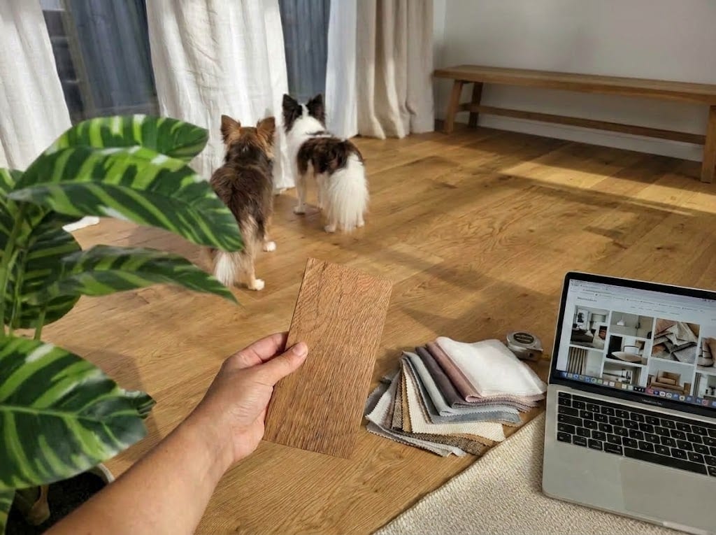 A hand holding a wood sample over an oak floor, beside fabric swatches and a laptop, while two small dogs look out of the window.